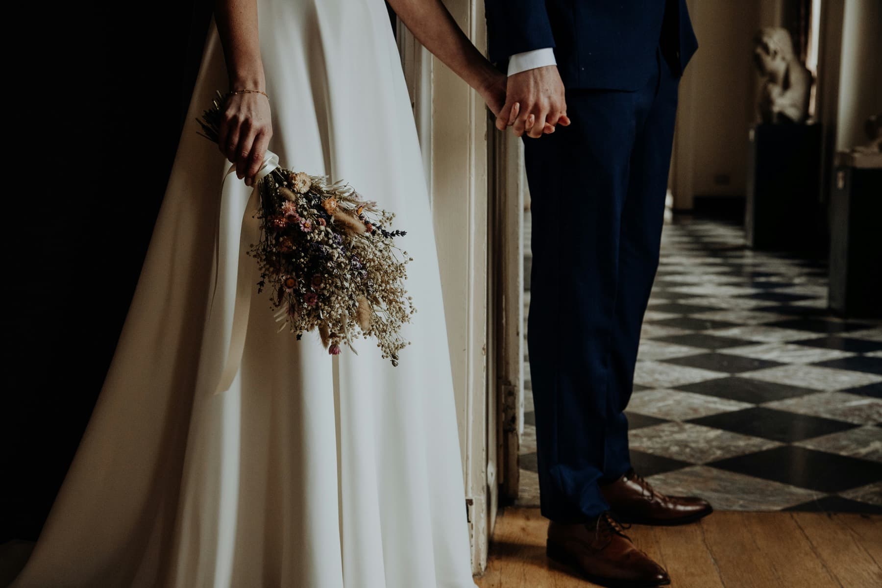 A couple holding hands in a hallway, her ivory dress and dried bouquet beside his navy suit